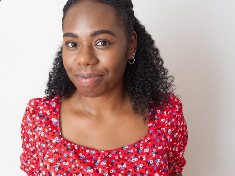 Smiling headshot of a dark skinned woman wearing a flowery red top against a light background, shot by a kent headshot photographer
